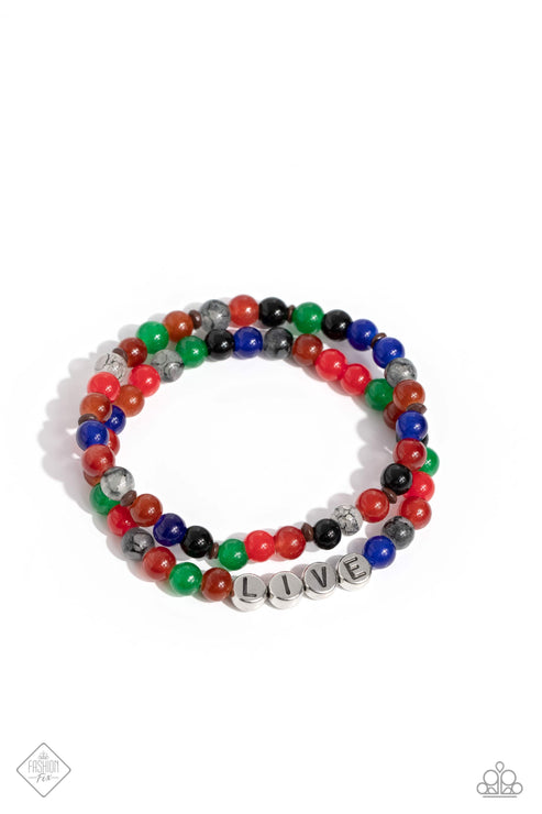 Countless stone beads in a range of vibrant colors are threaded along a pair of stretchy bands to create a stack of earthy bracelets. Silver discs stamped with black letters are centered within one of the colorful strands, spelling out the word, 