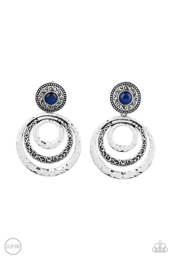 A glowing blue cat's eye stone is pressed into a studded silver frame embossed in swirling filigree. Varying in hammered and embossed textures, three silver rings ripple out from the bottom of the cat's eye frame for a whimsical finish. Earring attaches to a standard clip-on fitting.  Sold as one pair of clip-on earrings.