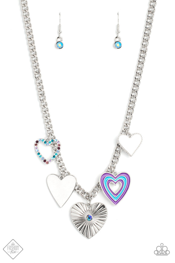 A collection of whimsical heart charms, each with its own unique features, gathers along a flat, silver curb chain. One heart is bordered in tiny silver studs, another is painted in bands of blue and purple, a third heart radiates with crimped texture and is topped with an iridescent rhinestone center, another charm radiates with its high-sheen finish, and lastly, a heart silhouette is bordered in tiny rhinestones in shades of blue and purple. 